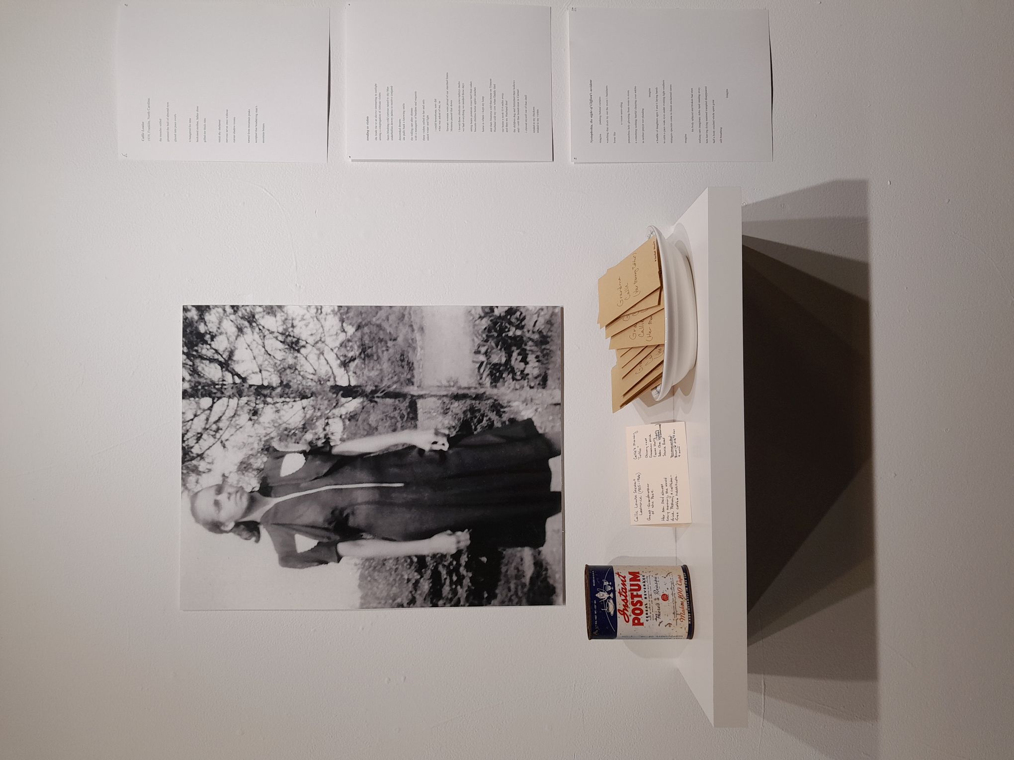 A black and white image of a woman wearing a dress and standing in a forest hangs on a wall. To the right of the image are print-outs of poems hung vertically. In front of the image is a shelf with a Postum can, a notecard, and tea bags in a bowl.