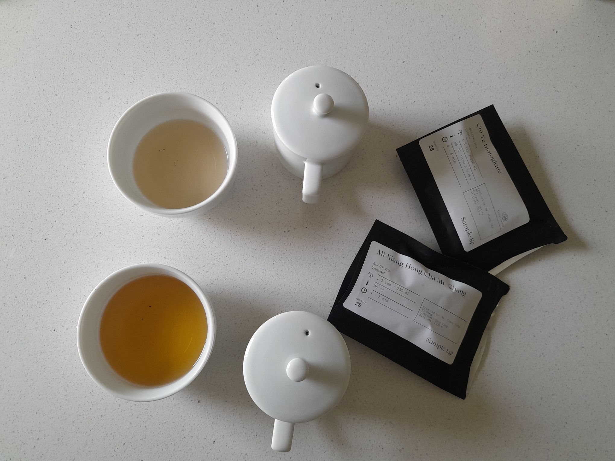 Two white tea bowls containing a light orange liquid and a dark orange liquid, two tea cupping containers, and two tea packages.